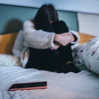 trauriges Mädchen sitzt auf dem Bett, im Vordergrund ein Handy