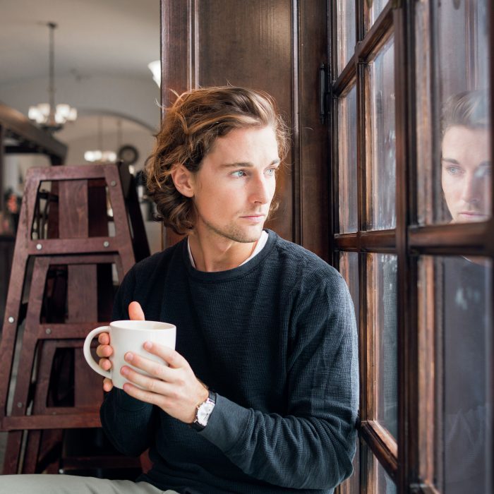 junger Mann sitzt alleine mit einem Becher in der Hand in einem Café und schaut nach draußen, sein Gesicht spiegelt sich im Fenster
